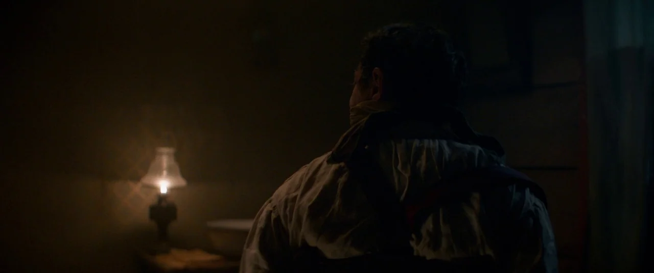 The Sisters Brothers (2018) movie still: medium — A dark, dimly lit room contains a small table holding a lit oil lamp. A man sits with his back to…