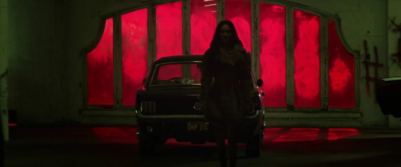 Zeroville (2019) movie still: fullbody — A dark, industrial interior space features a large arched window backlit by bright red light. A woman in a…