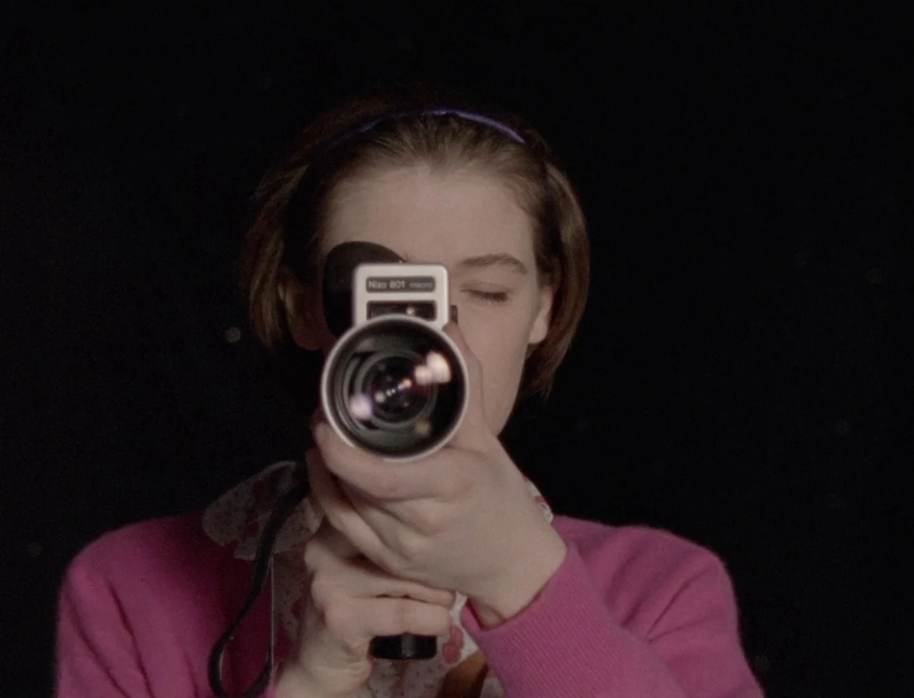 The Souvenir: Part II (2021) movie still: closeup — A person stands against a black background holding a vintage super 8 movie camera. The subject…