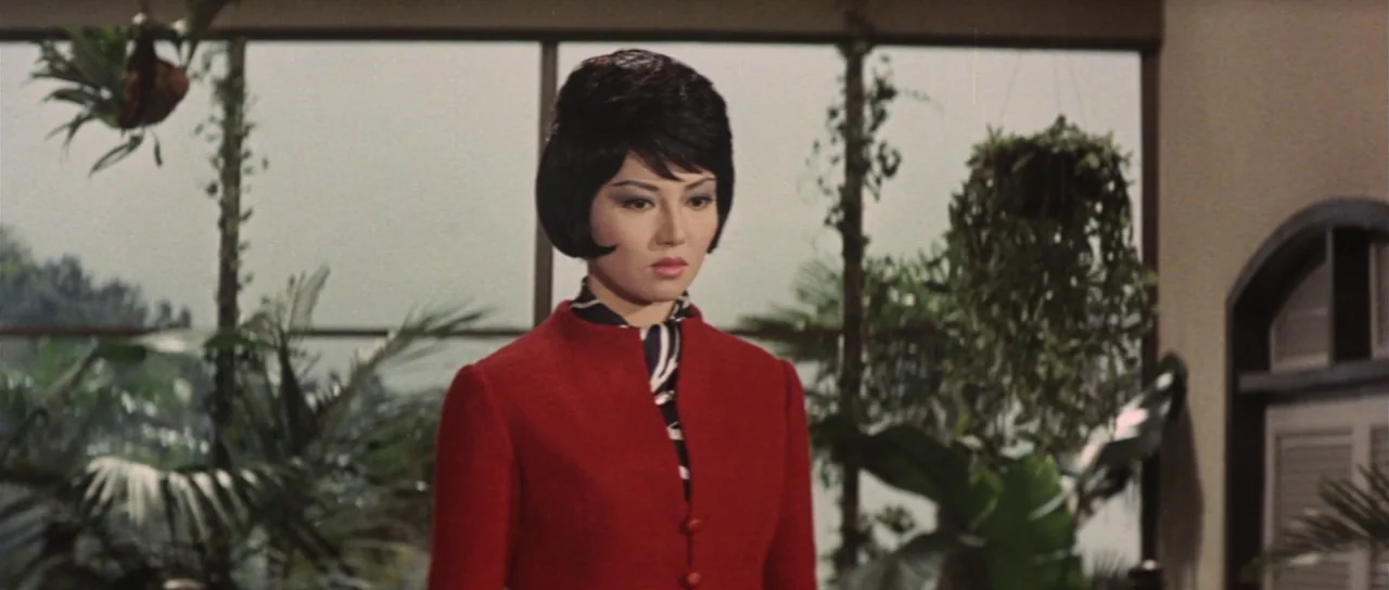 Invasion of Astro Monster (1965) movie still: medium — A woman stands in a sunroom filled with various potted plants and large windows. The interior…