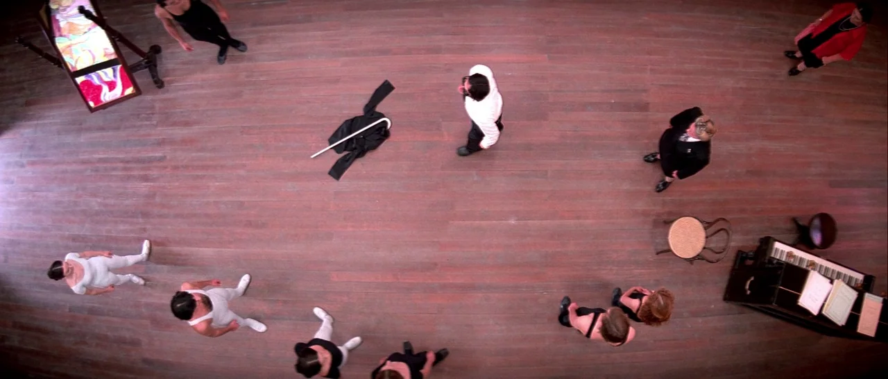Suspiria (1977) movie still: wide — A spacious room features a polished wooden floor and a corner piano. Multiple individuals stand in an arranged…