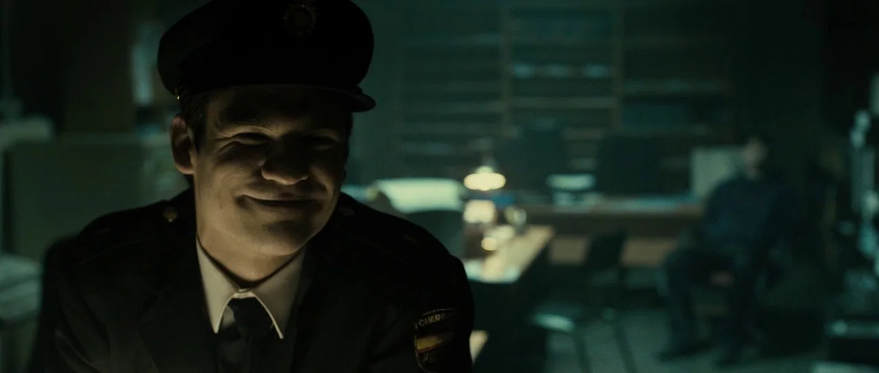 The Imposter (2012) movie still: closeup — A darkened, dimly lit room contains a desk and blurred figures in the background. A man in a uniform…