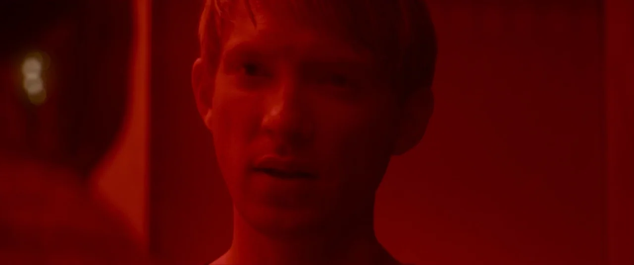 Ex Machina (2015) movie still: closeup — A dark indoor space is lit entirely by a deep red light source. A young man stands in the foreground facing…
