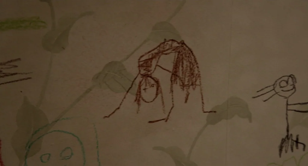 Mama (2013) movie still: closeup — A cream-colored wall is decorated with faint floral wallpaper and several crayon drawings. One sketch depicts a…