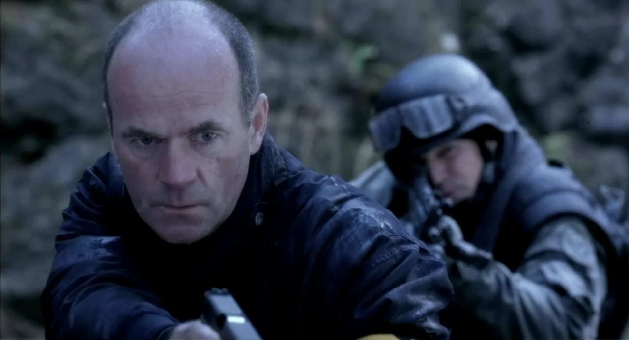 The Hunted (2003) movie still: medium — A rugged exterior location with a textured stone wall in the background. A man holds a pistol forward while…