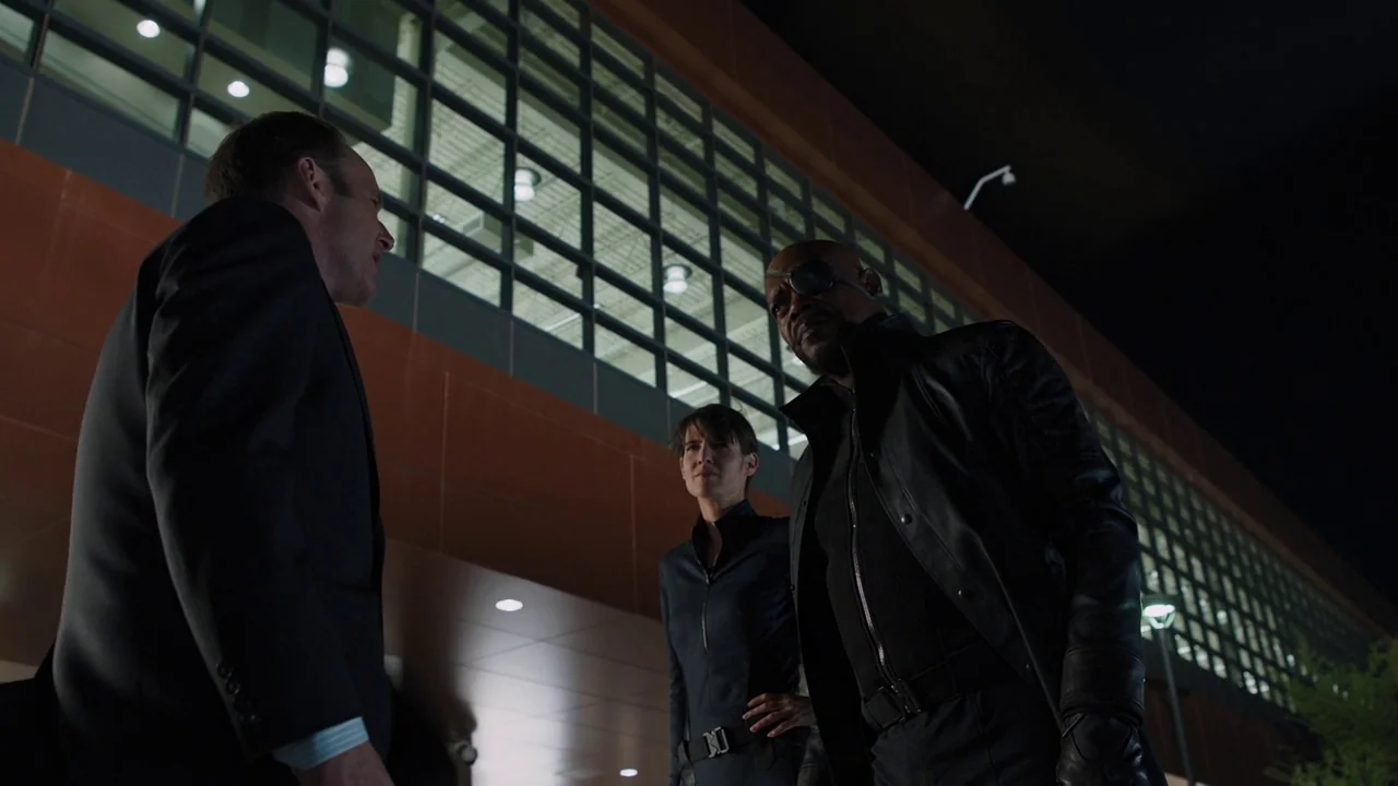 The Avengers (2012) movie still: medium — A dark night exterior features a modern office building with large grid glass windows. Three individuals…