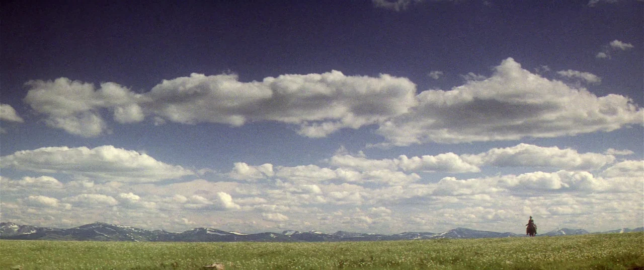 Heaven’s Gate (1980) movie still: wide — A vast, green grassy prairie stretches beneath a expansive sky filled with white clouds. A solitary figure…