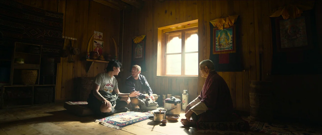 The Monk and The Gun (2023) movie still: wide — A wooden cabin with walls made of horizontal planks and various religious wall hangings. Three men…