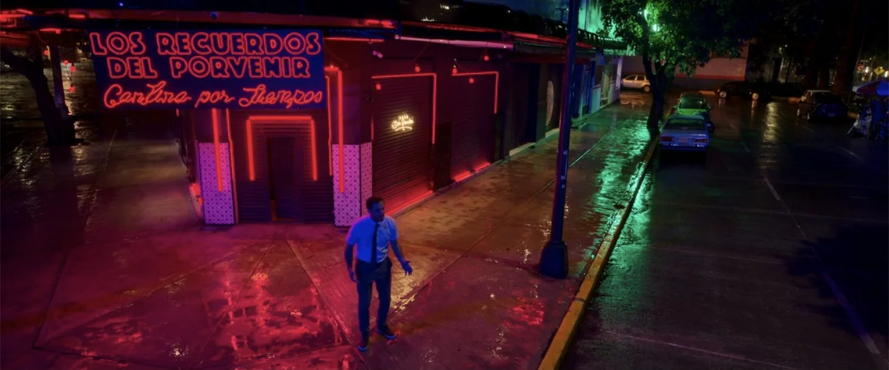 Tequila Re-Pasado (2023) movie still: wide — A nighttime urban street corner features a storefront with bright red neon Spanish signage. A man in a…