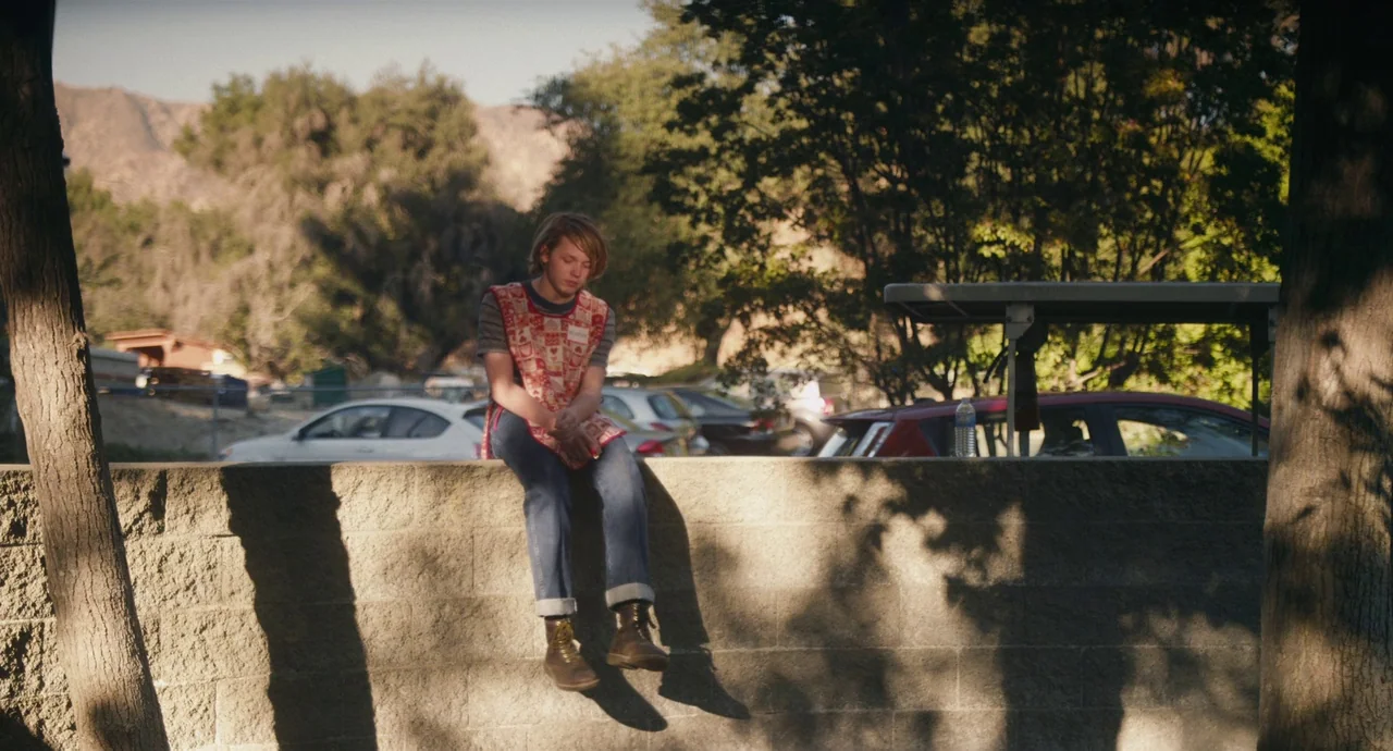 Palo Alto (2013) movie still: fullbody — A teenage boy sits on a low concrete wall in a sunlit parking lot area. Cars are parked in the background…