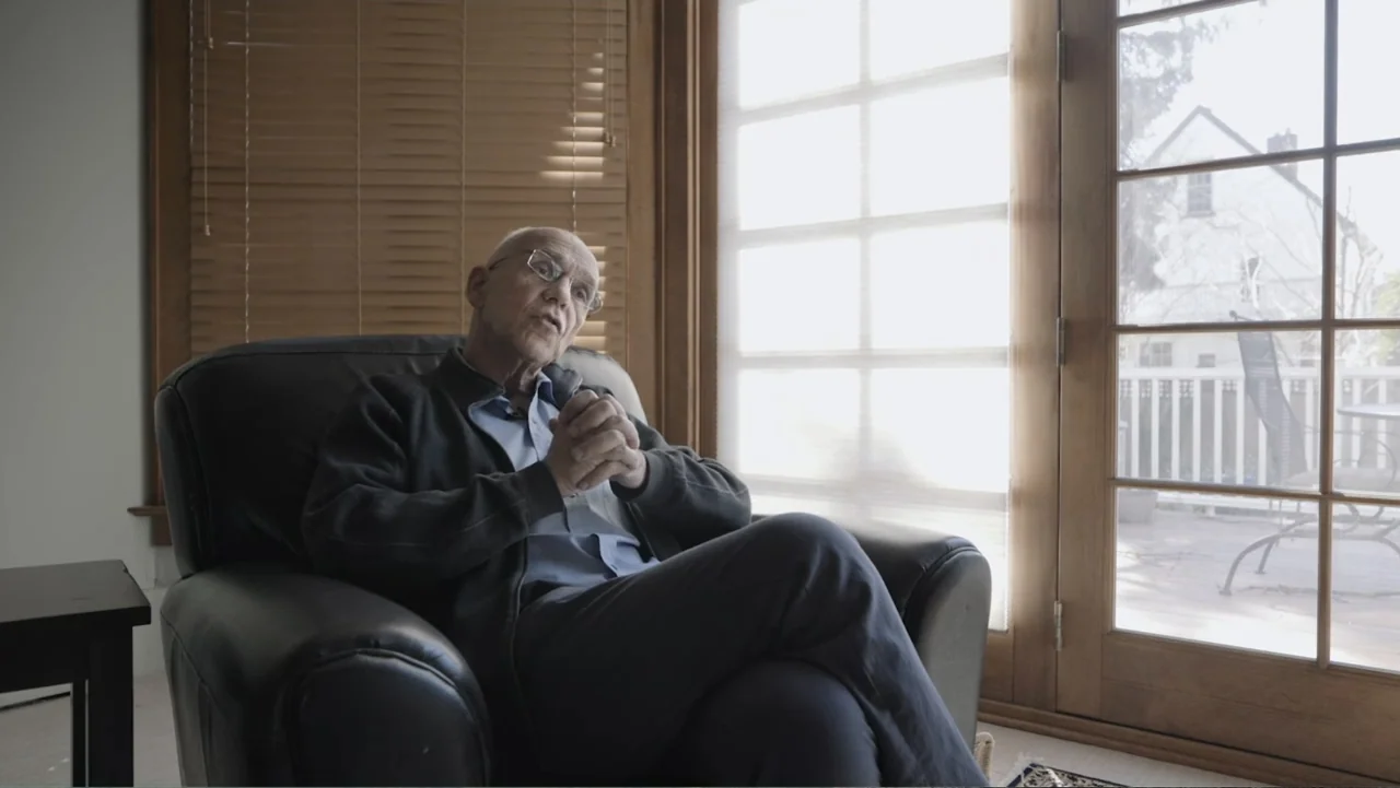A Gift from God (2019) movie still: medium — An interior living room with a large window and wooden blinds. An elderly man sits in a black leather…