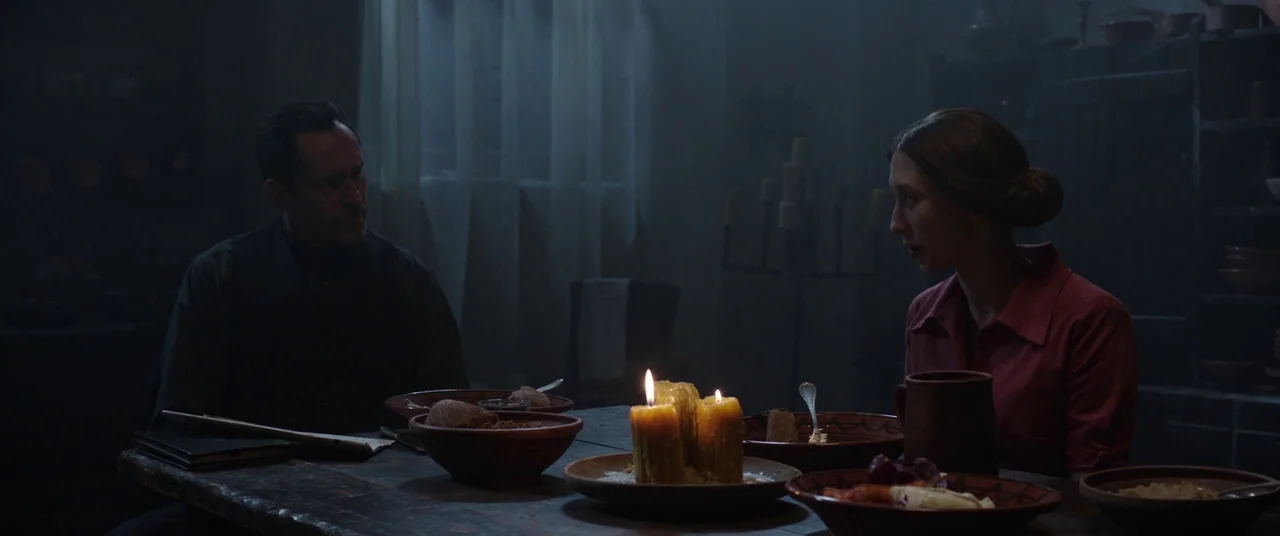 The Nun (2018) movie still: medium — A dimly lit room contains a rustic wooden table set with simple bowls and candles. A man and a woman sit facing…