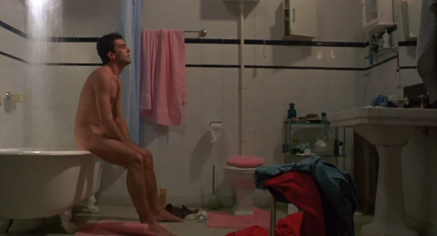 Matador (1986) movie still: fullbody — A tiled bathroom with pink accessories and an open shower head above a tub. A nude man sits on the side of…