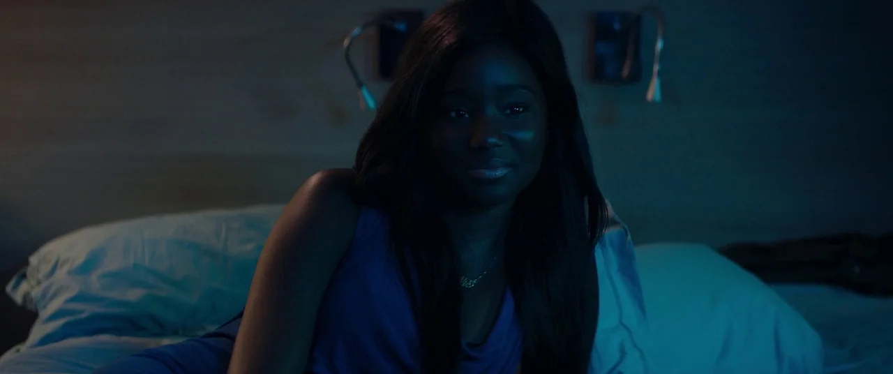 Girlhood (2014) movie still: medium — A dark, dimly lit bedroom at night with two wall-mounted light fixtures behind the subject. A young woman with…