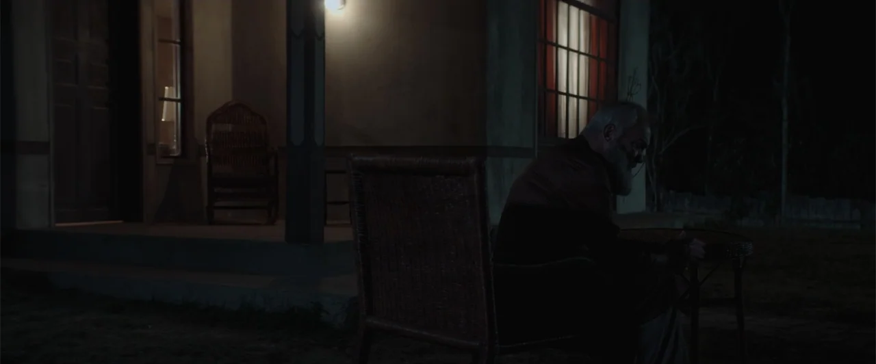 My Neighbor Adolf (2022) movie still: medium — A dark, nighttime exterior of a home's porch with wooden pillars and wicker furniture. An older man…