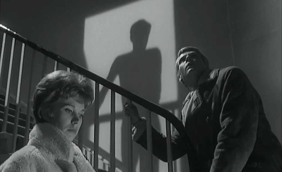 The Servant (1963) movie still: medium — An interior staircase features a wooden railing and a tall wall with a shadow. A man and woman stand on the…