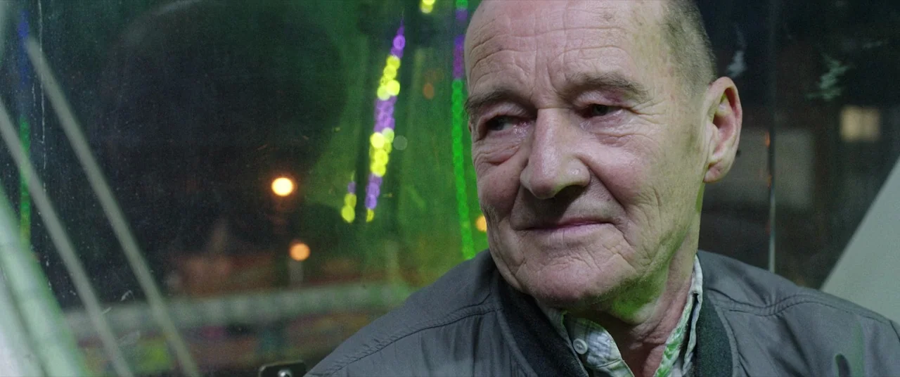 Bull (2021) movie still: closeup — A man sits inside a glass-enclosed amusement ride at night. Blurred green and yellow lights from the carnival…