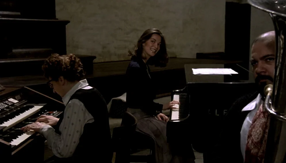 Orchestra Rehearsal (1978) movie still: medium — A dimly lit rehearsal room contains an organ, a grand piano, and a tuba player. Three musicians…
