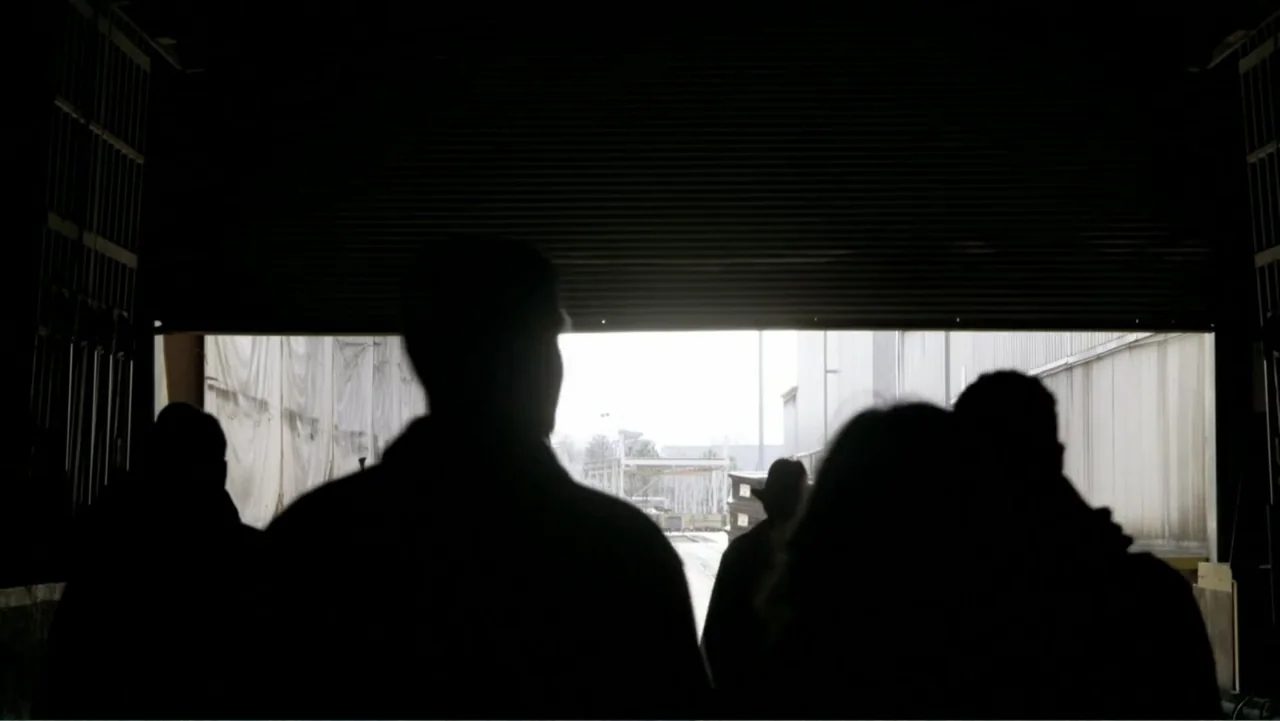 Ben 10: Race Against Time (2008) movie still: medium — A dark industrial warehouse interior looks out toward an open loading bay. Three individuals…