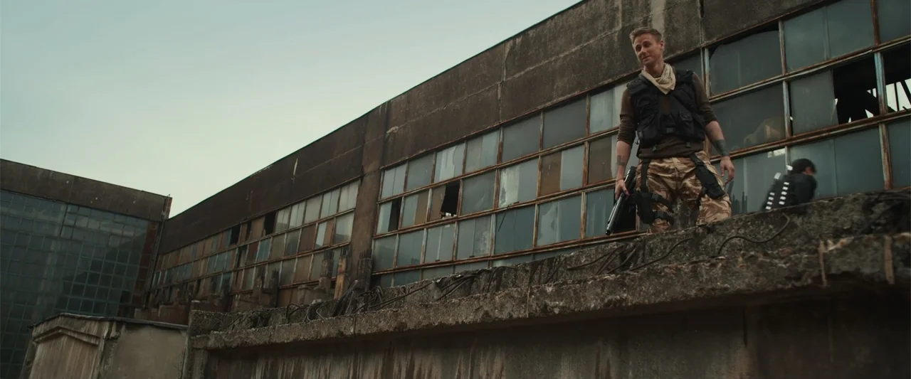 The Condemned 2 (2015) movie still: fullbody — A man stands atop a weathered concrete building with multi-paned glass windows. He holds a shotgun…
