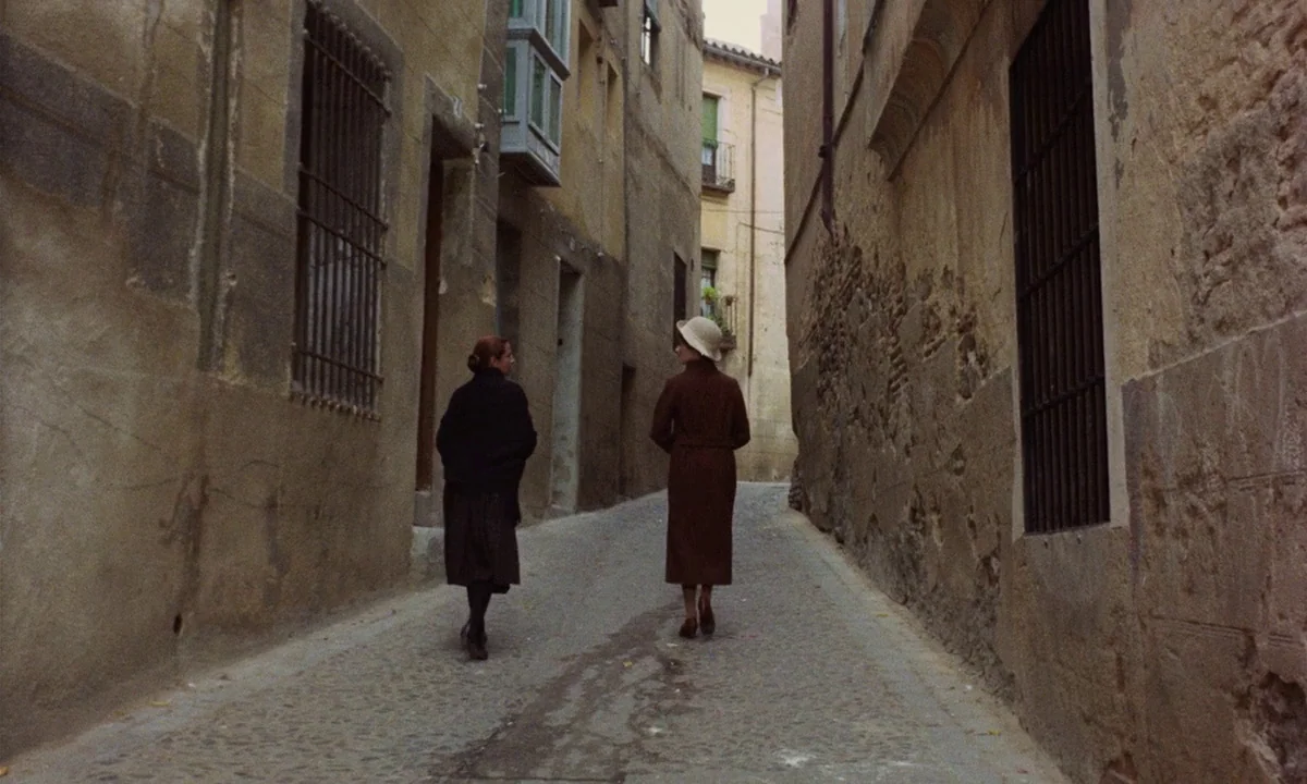 Tristana (1970) movie still: fullbody — A narrow cobblestone alley lined with tall, aged stone buildings and iron-barred windows. Two women in dark…