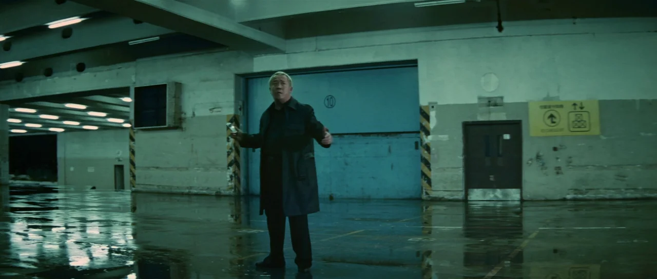 Infernal Affairs (2002) movie still: fullbody — A wet, industrial loading dock has bare concrete walls and high, recessed lighting strips. A…