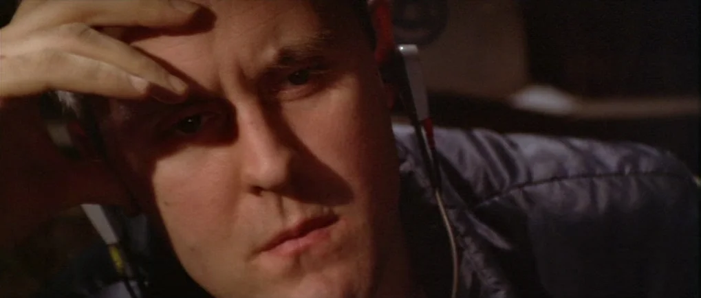 Blow Out (1981) movie still: closeup — A dark, cramped interior space with minimal ambient light. A man wearing wired headphones gazes forward while…