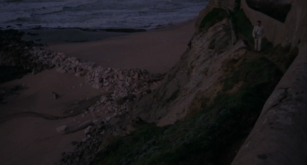 Anatomy of Hell (2004) movie still: wide — A rugged cliff overlooks a sandy beach and dark ocean waves at dusk. A man walks along a narrow dirt path…
