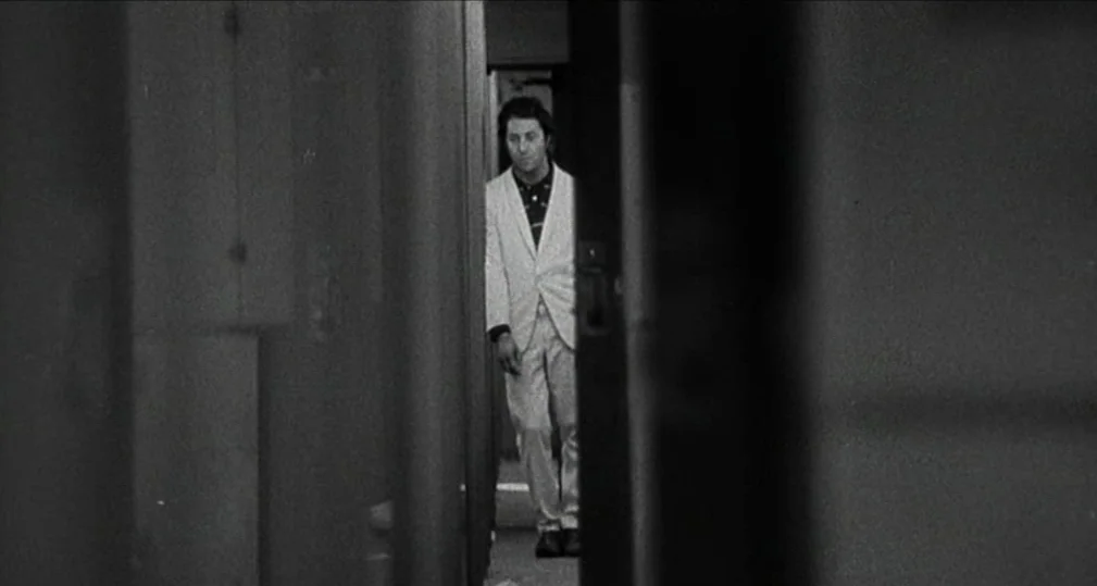 Midnight Cowboy (1969) movie still: medium — A man stands within a narrow corridor viewed through a dark, shadowed doorway. The hallway is sparsely…