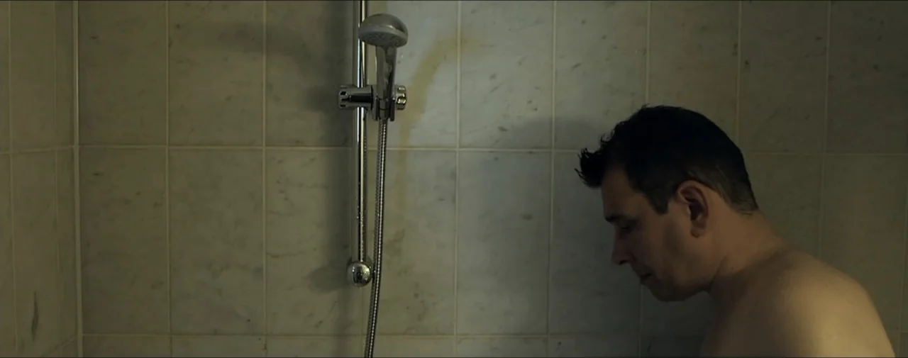 Hunting for Hedonia (2019) movie still: medium — A shower stall with light-colored marble tiles and a chrome shower fixture. A man stands partially…
