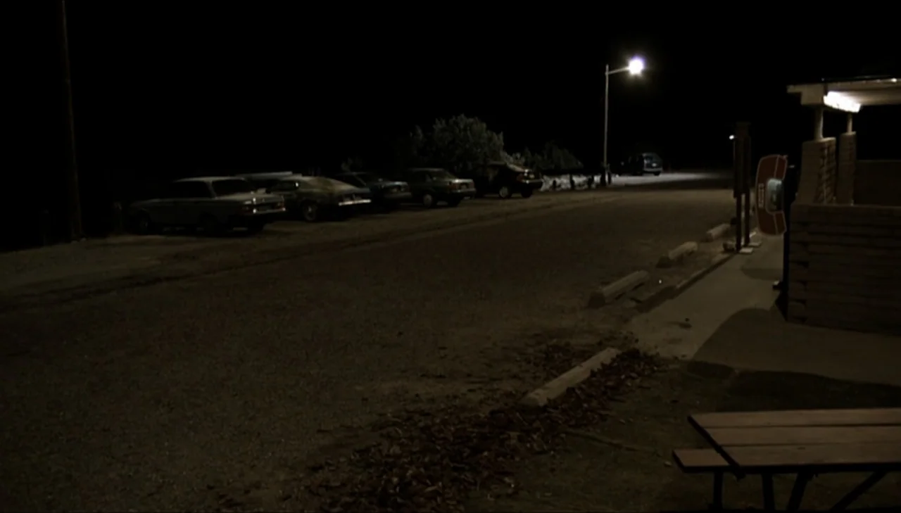 Dark Country (2009) movie still: wide — An empty gravel parking lot at night is illuminated by a single streetlamp. Several parked cars line the…
