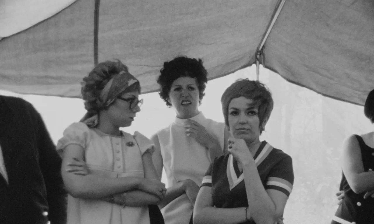 Multiple Maniacs (1970) movie still: medium — Three women stand beneath a fabric canopy during an outdoor daytime event. They are clustered together…