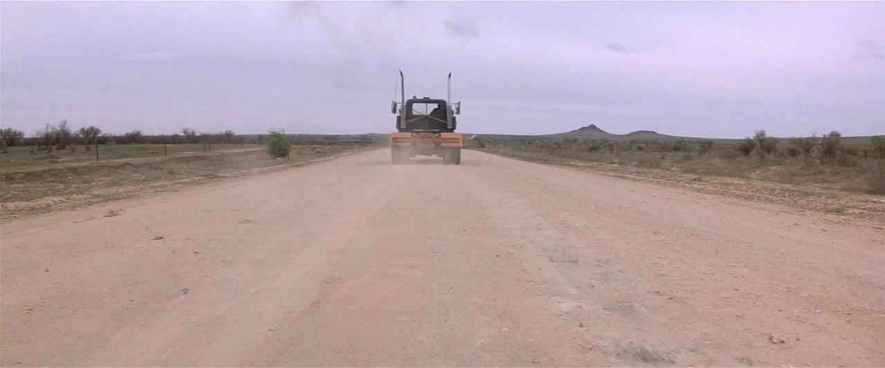 Mad Max 2 (1981) movie still: wide — A wide dirt road stretches through an expansive, arid landscape under an overcast sky. A large industrial truck…