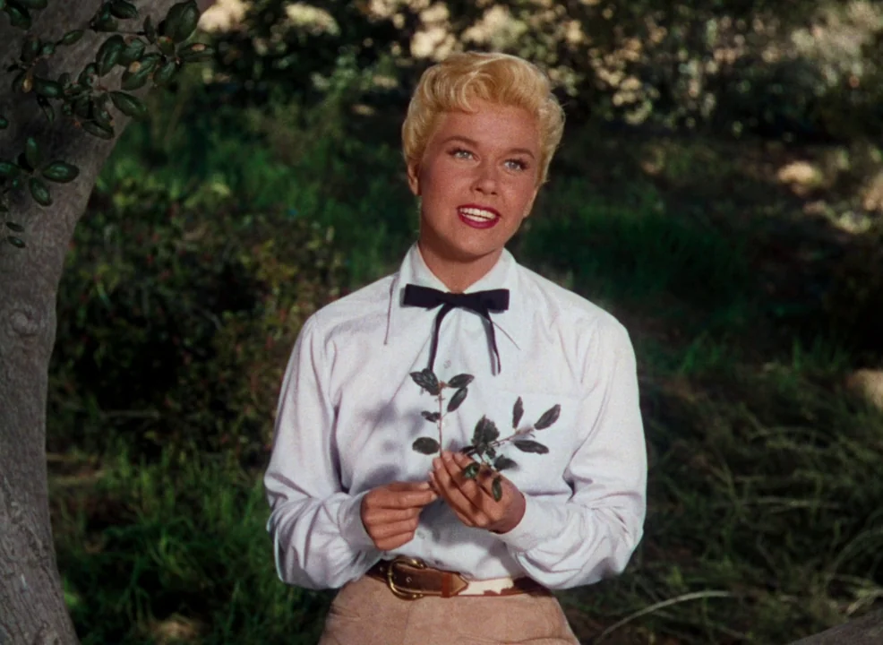 Calamity Jane (1953) movie still: medium — A woman stands in a lush, green wooded area with dense surrounding foliage. The woman holds a small sprig…