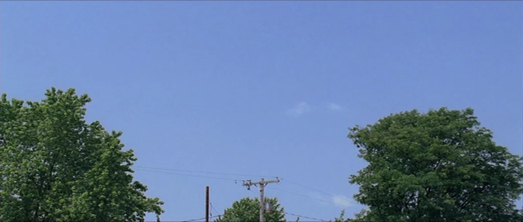 Take Shelter (2011) movie still: establishing — A clear blue sky extends above dense green trees flanking a utility pole. The pole stands centrally…