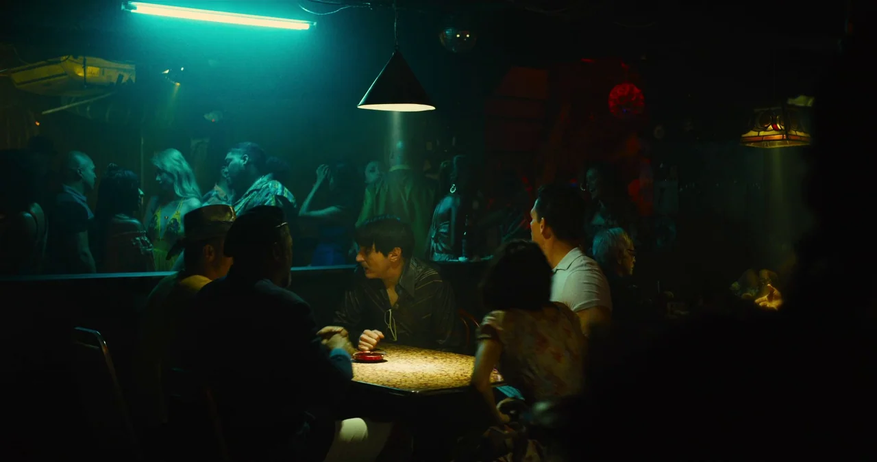 The Suicide Squad (2021) movie still: medium — A dimly lit bar interior filled with people in a low-key, colorful environment. A small group sits at…