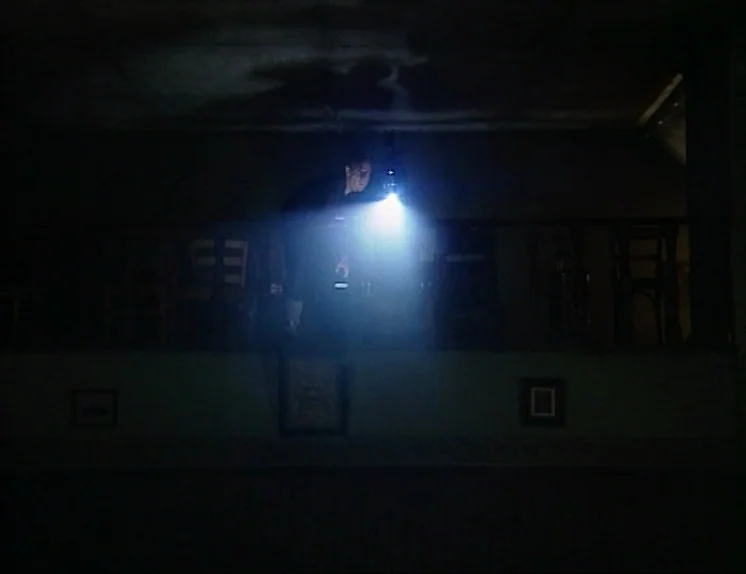 In The Presence of a Clown (1997) movie still: medium — A dark balcony interior features a wooden railing with chairs tucked along the back. A man…
