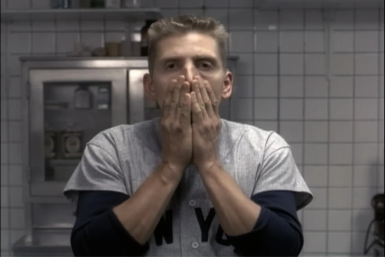 Unification of Japan 61 (2024) movie still: medium — A white tiled room with a metallic cabinet in the background. A man wears a baseball jersey…