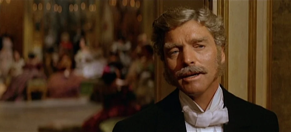 The Leopard (1963) movie still: medium — A crowded, dimly lit 19th-century ballroom with guests blurred in the deep background. An older man in…