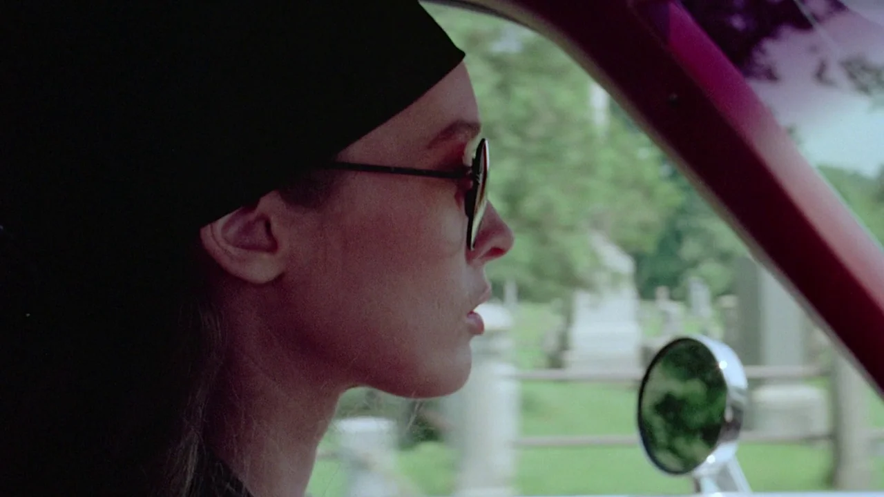 I Spit On Your Grave (1978) movie still: closeup — A woman drives a vehicle past a graveyard with white headstones visible through windows. The…