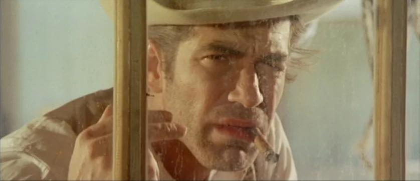 Django Kill (1967) movie still: closeup — A man in a tan hat is framed behind a dusty, multi-pane window. He stares directly ahead with a cigar held…