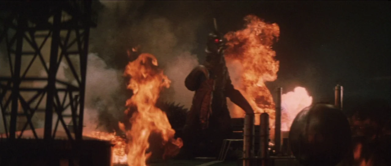 Godzilla Vs Gigan (1972) movie still: wide — An industrial facility at night is covered in large, bright flames and thick smoke. A large mechanical…