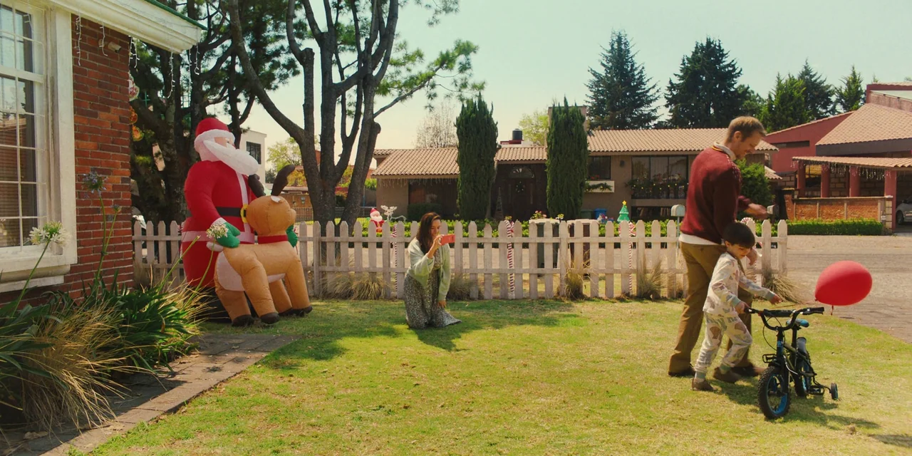 Silent Night (2023) movie still: wide — A residential front yard features a white picket fence decorated with Christmas candy canes. A man assists a…