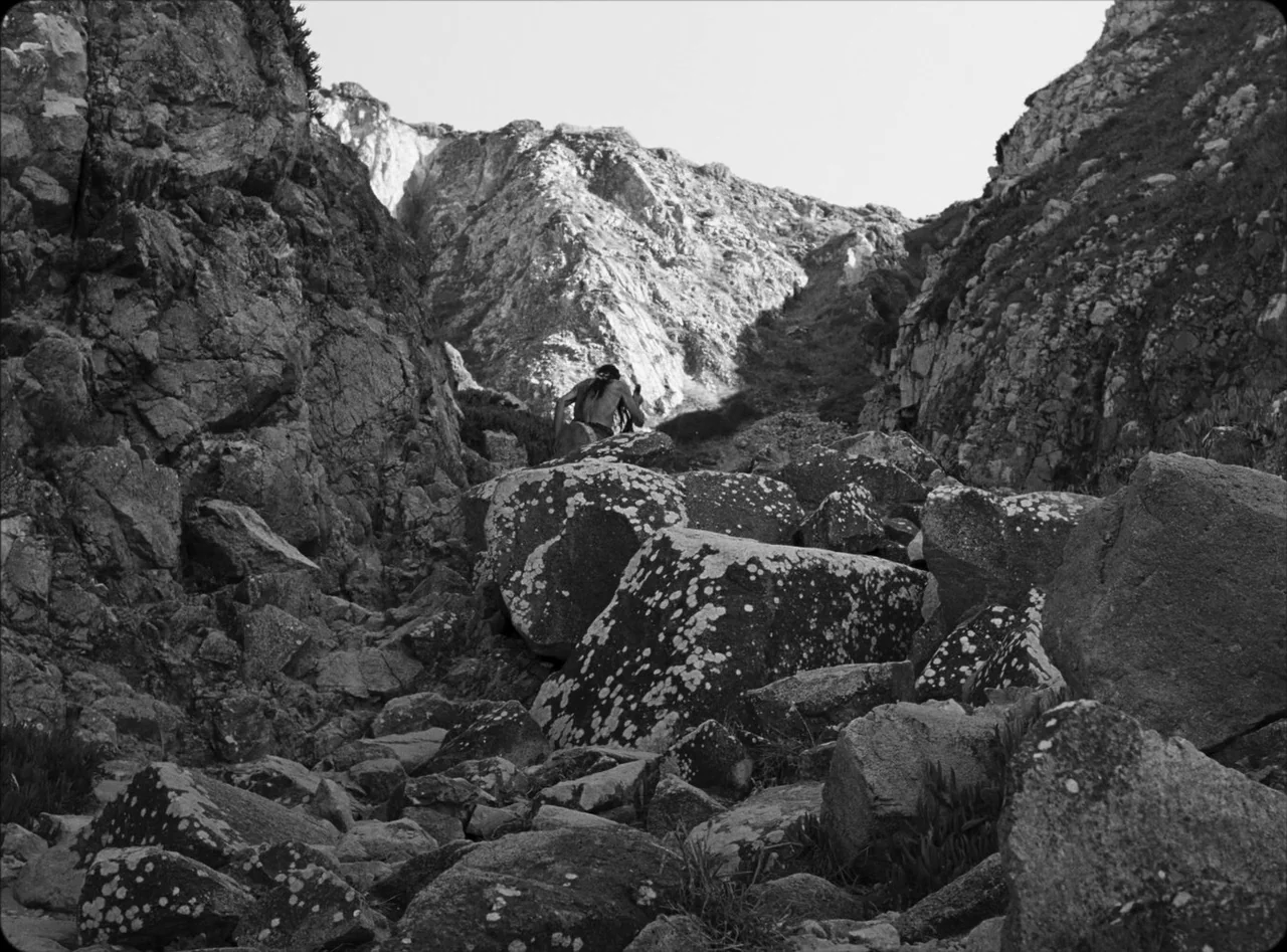 Eureka (2024) movie still: wide — A rugged rocky gully is lined with massive, lichen-covered boulders leading toward a slope. A solitary figure with…