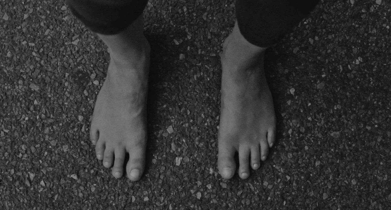 Frances Ha (2012) movie still: closeup — A pair of human feet stands directly upon a coarse, dark asphalt ground surface. The individual is dressed…