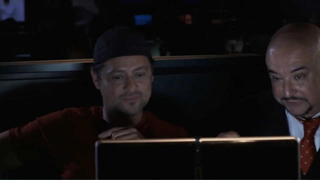 Hello I Love You (2018) movie still: medium — A dimly lit room with dark partitions where two men sit side by side. The individuals look downward at…