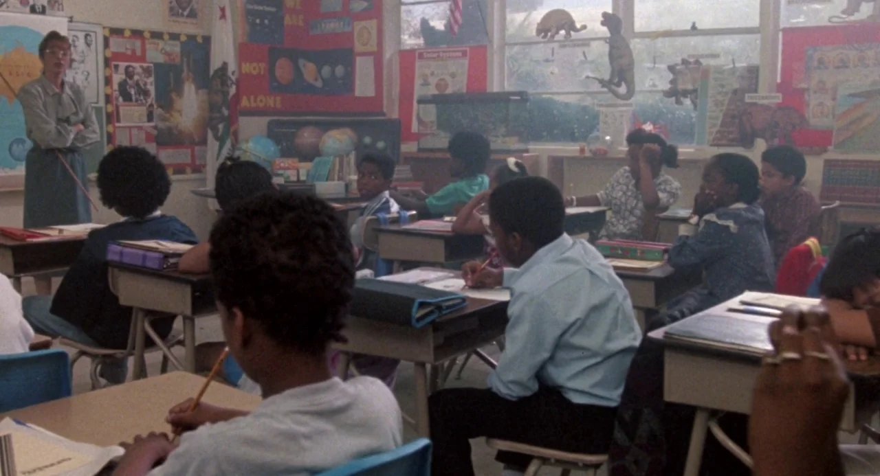 Boyz In The Hood (1991) movie still: wide — An elementary school classroom is filled with children seated at wooden desks facing forward. A teacher…