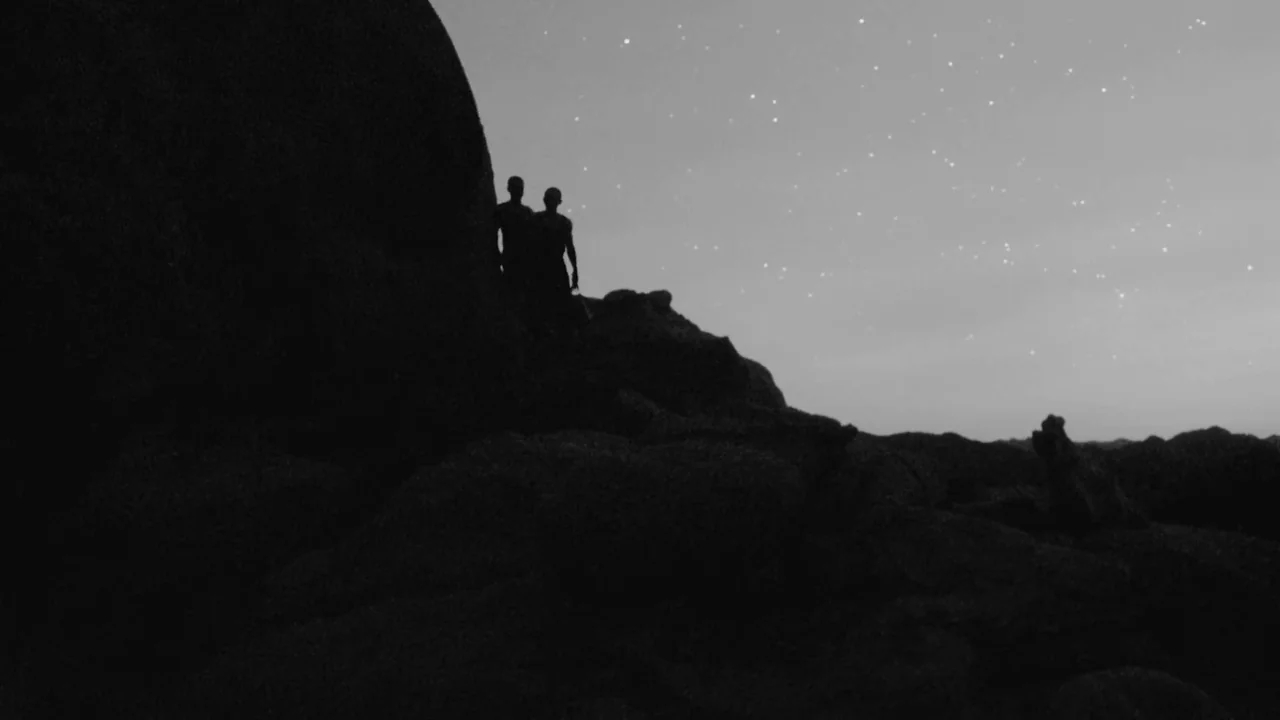 Divinity (2023) movie still: wide — A dark rocky landscape under a clear night sky filled with faint stars. Two people stand close together on top…