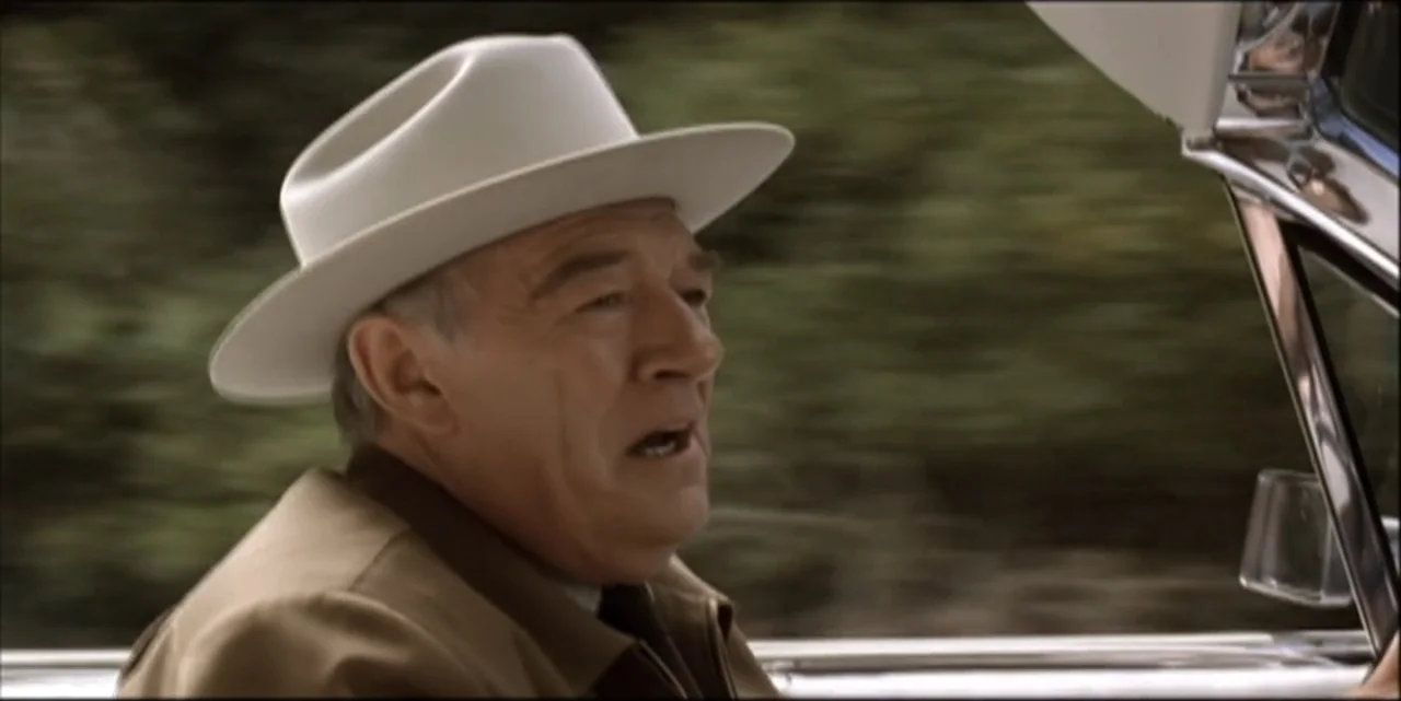 Path to War (2003) movie still: medium — An older man drives a vehicle through a rural area with motion blur visible. The man wearing a cowboy hat…