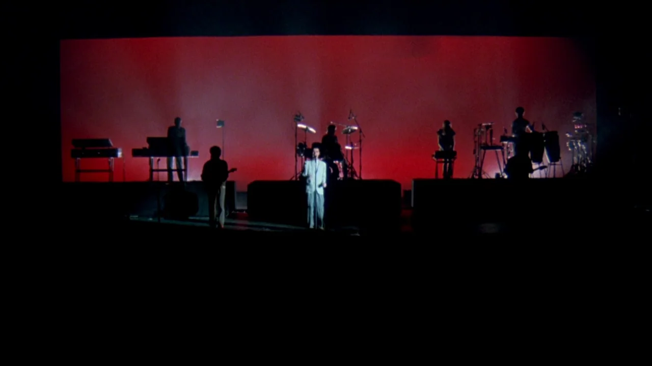 Stop Making Sense (1984) movie still: wide — A dark concert stage featuring a solid red background with multiple musicians in silhouette. The lead…